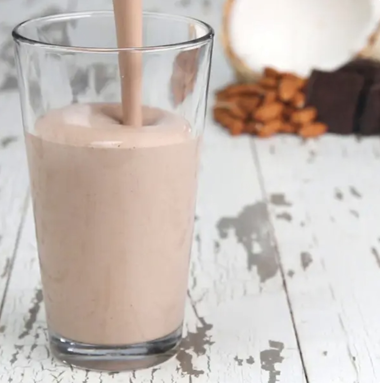 A glass is being filled with a chocolate milkshake. The background shows almonds, chocolate, and a coconut half