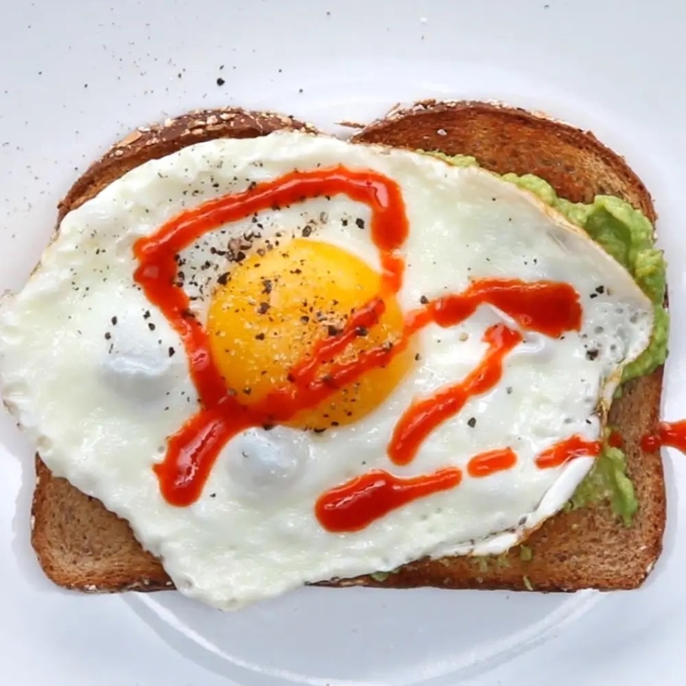 A piece of toast with avocado spread, topped with a sunny-side-up egg, drizzled with hot sauce, and sprinkled with black pepper