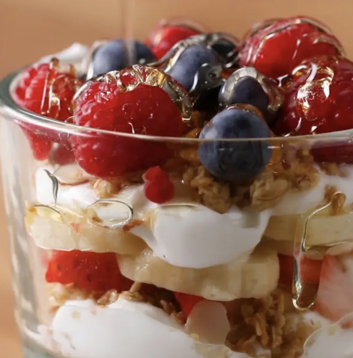 A close-up of a parfait layered with yogurt, granola, sliced bananas, strawberries, raspberries, blueberries, and honey drizzled on top