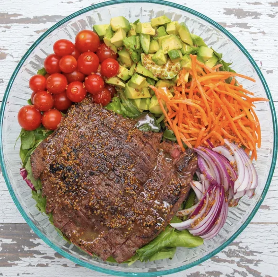 Glass bowl with grilled steak, cherry tomatoes, diced avocado, shredded carrots, and sliced red onions over a bed of lettuce