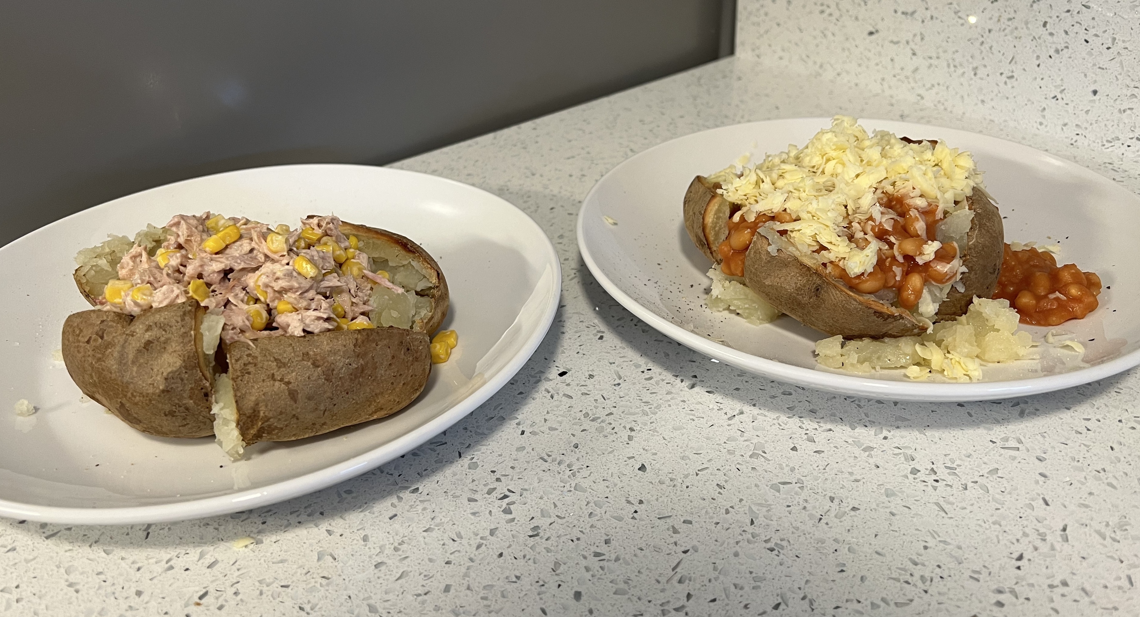 Two baked potatoes on plates, one topped with tuna and sweetcorn and the other with baked beans and shredded cheese, placed on a kitchen countertop