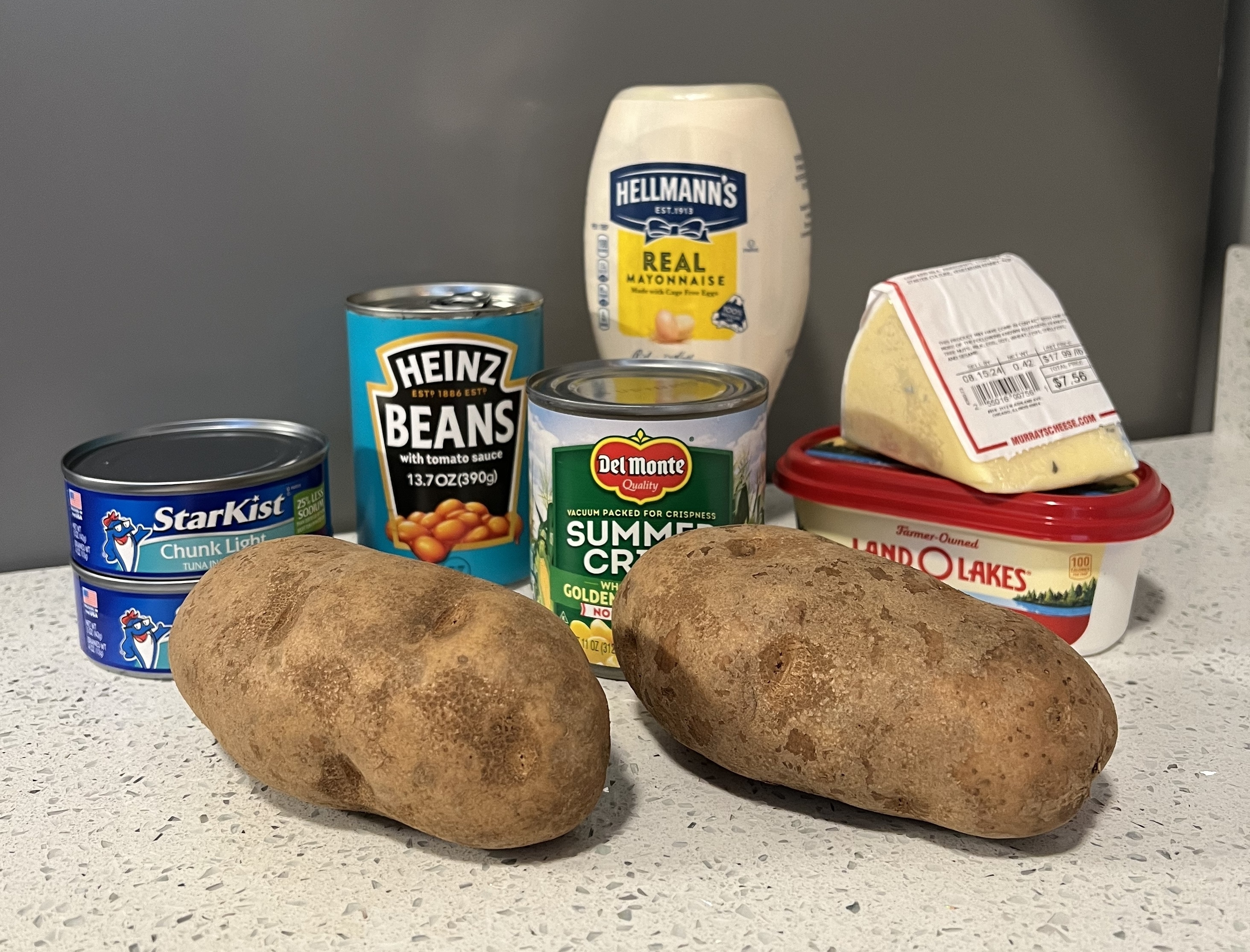 Two russet potatoes sit on a countertop surrounded by Heinz beans, Hellmann&#x27;s mayonnaise, Starkist tuna, Del Monte corn, cheese, and Land O&#x27;Lakes butter