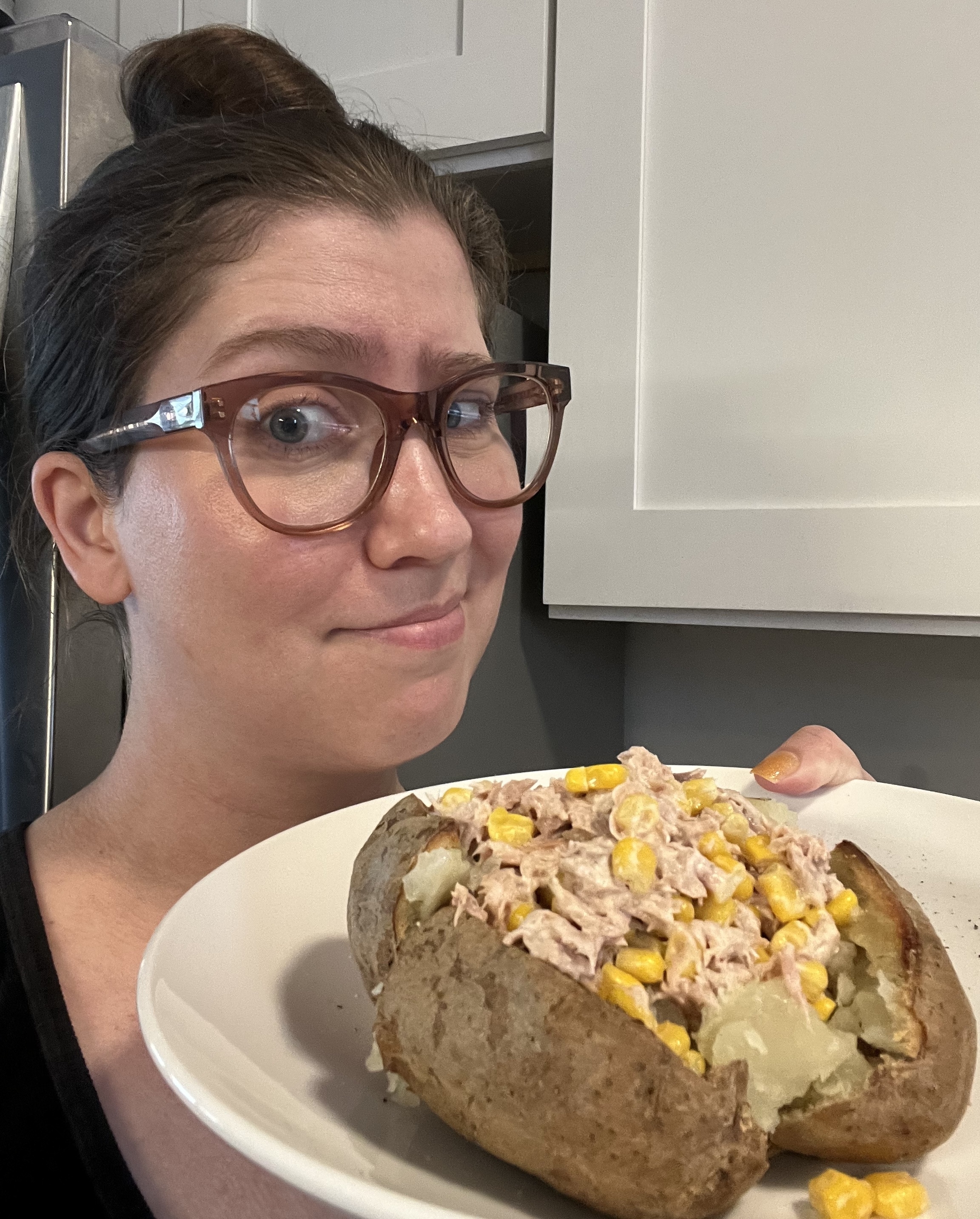 A woman wearing glasses holds a plate with a baked potato topped with tuna salad and corn