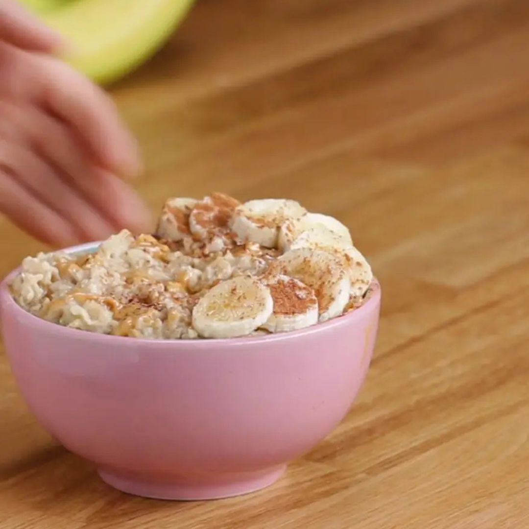 A pink bowl of oatmeal topped with sliced bananas, nuts, and a sprinkle of cinnamon sits on a wooden table. A hand is slightly blurred in the background