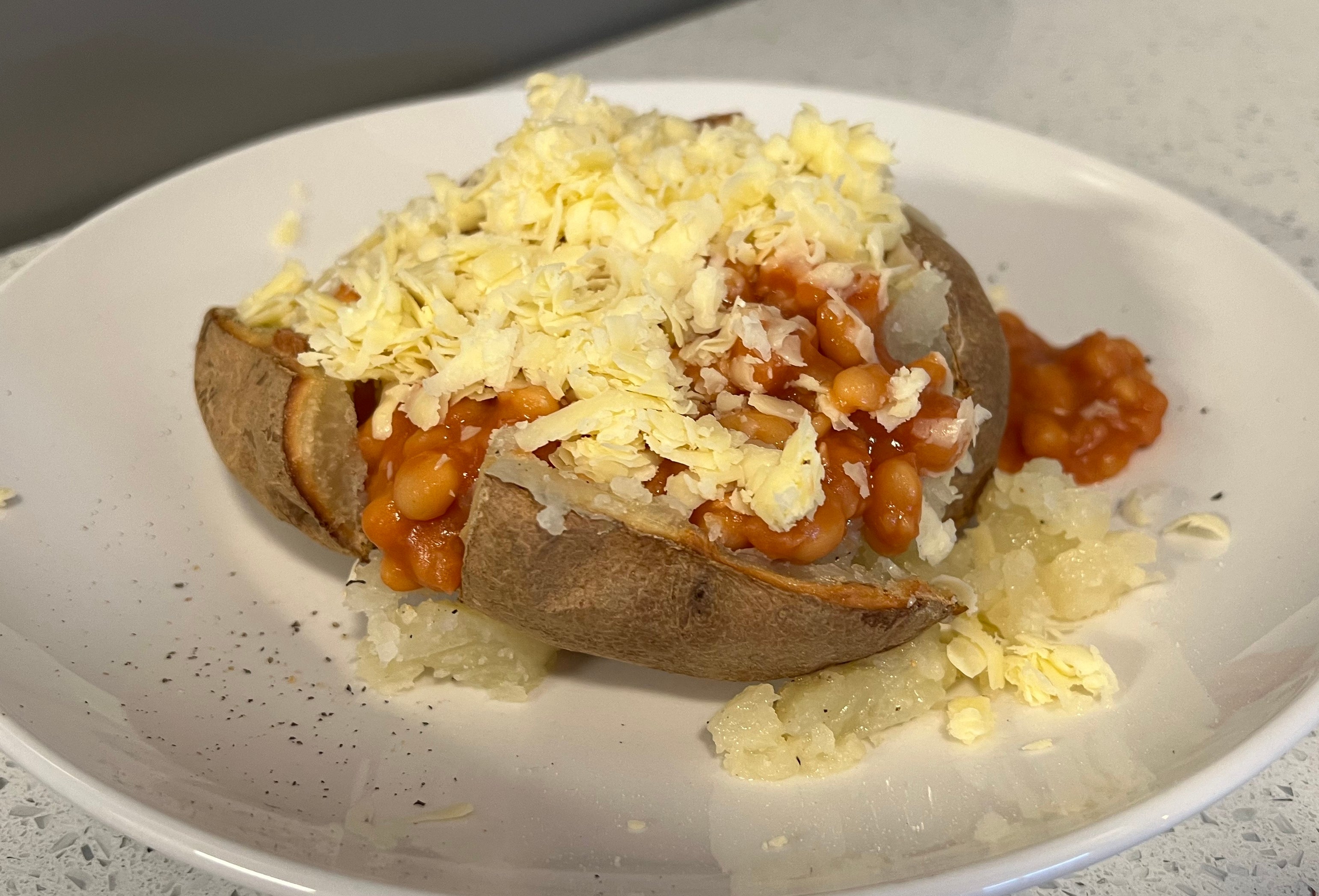 A baked potato topped with baked beans, grated cheese, and a sprinkle of black pepper, served on a white plate